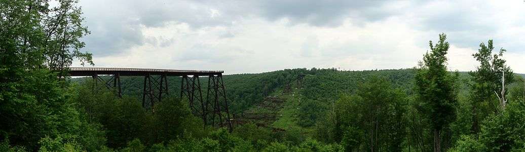 View of the remains of a bridge across a valley. Part of the bridge in foreground and a portion at its far end remain standing, whereas the rest is collapsed and lying on the valley floor.