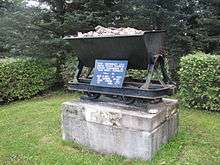 A monument to the site where the first tonnes of oil shale were mined in Estonia. A metal hopper containing rock is mounted above a concrete block about one metre wide and one-half metre deep. A blue plaque with white lettering in the Estonian language explains its significance.