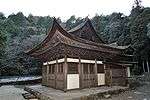 Small wooden building with white walls and hip-and-gable roof connected to another wooden structure in the back.