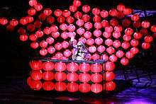 A woman, wearing sunglasses, sits on a table and waves her hand. The table and her background is covered with small red lanterns.