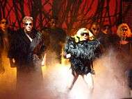 A group of people stand on a stage in black, feathery dresses and black sunglasses. Prominent among them is a blond woman, with a mouthpiece attached to her ear. Behind the group, a red background can be seen, interspersed with black thorn-like structures.