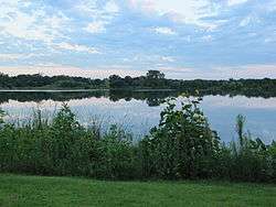 A placid lake lies under a pink/blue afternoon sky. In the foreground is a well groomed lawn and a border of plants, including a sunflower, that encircles that lake.