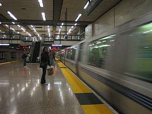 A near-empty station platform of an underground train station, with an escalator in the background. A train is leaving the station