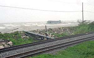 Rough waves on a lake. A railroad is situated along the bottom of the image on an embankment. A fishing hut is present near the lake.