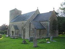 Gray stone building with square tower at left hand end. In the foreground are gravestones on grass.