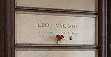 A white marble gravestone on the wall of a chapel, with only the name and dates of birth and death inscribed, a small photograph of the deceased and a bunch of red carnations