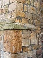 Orange to light brown irregular weathered stones forming a corner of a building, two of the stones with filled in rectangular central holes