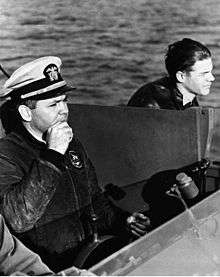 Three men at the con of a boat. One wears a white peaked cap and holds the wheel.