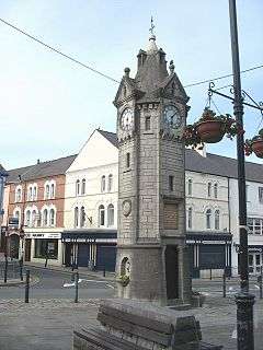 A tower-like structure in stone with a clock face on each side.  Above each clock is a gable and at the summit of the structure is a spire.