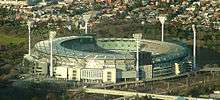 Aerial view of outdoor sport stadium. Erected around the stadium are six tall flashlights. The stadium are surrounded by houses, parklands and railway lines.