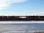 A frozen river passes through flat country. Short trees grow on the riverbanks; tall mountains are in the far distance.