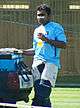 A man in a blue T-shirt, wearing white tracksuit trousers with pads. A grass-cutting machine can be seen in background.