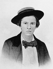 A young North American boy of the 19th century. In this black-and-white photograph, the boy looks into the camera nervously, a straw boater perched upon his head. He wears a white shirt, dark jacket and a large, dark-coloured bow tie.