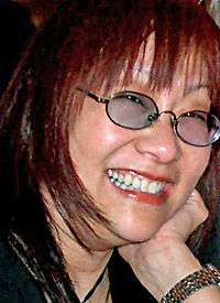 Profile picture of a bespectacled Asian woman in her early fifties. She has long red hair, and shows a toothy smile.