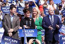 Todd Palin, Sarah Palin (behind a podium), Cindy McCain, John McCain together on an outdoor stage during daytime, crowd holding blue-and-white "McCain Palin" signs around them