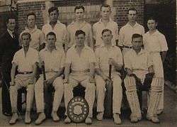 A group of 11 young cricketers in an official photo. They are wearing their white cricket uniforms, and five sit in the front row, and six stand behind them, along with a middle-aged man, their coach, in a dark suit and tie. Some are holding bats and wearing pads and a shield is bat the foot of the central boy in the front row.