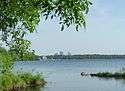 A bandshell and skyscrapers are visible in the distance across a slightly choppy lake, seen past small spits of grass-covered land that extend from either side of the frame.