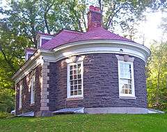 A stone building with red metal roof and a rounded projection in front. It is sheltered by trees to the rear.