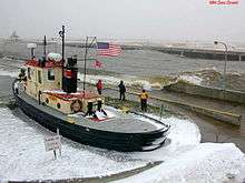 Tugboat at dockside, with three workers watching waves break over a sea wall