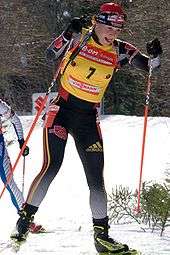 A woman cross country skis uphill towards the camera, holding a red ski pole in each hand. She wears black winter sportswear, a red cap and a yellow jersey with the number 7. A second skier behind her can be seen on the left.