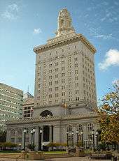 A pale yellow building with a clock tower on top stands behind a plaza with a lawn and street light poles