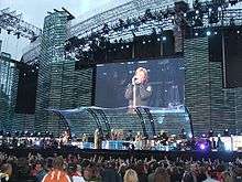 Full view of four men performing on stage which is lit by aqua-coloured lights. The most central man, dressed in mostly black, stands with his left arm reached over his head and his right arm holding out a microphone stand towards the front of the stage. A second man wearing a black tank top and maroon pants stands to the left playing his black and white guitar. Behind him is the third man with long, curly, blonde hair, suiting all black playing the keyboard. In the upper right corner is the upper body of a fourth man behind a drum kit.