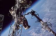 A man dressed in a spacesuit clings to a truss structure, manipulating a second truss with his left hand. Cabling can be seen running along both trusses, and the Earth's horizon can be seen in the background against the blackness of space.