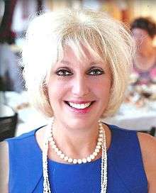 A photograph of a smiling, blond-haired, brown-eyed, middle-aged woman in a blue blouse.