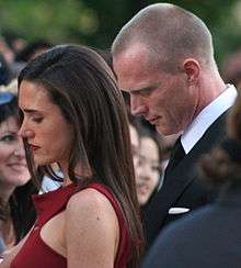 A brown hair woman signs autographs for fans. She wears a red dress. Behind her there is a blond man dressed with a suit. The woman and the man are facing a crowd of fans.