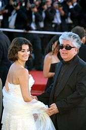 In the photo a Caucasian male and a Hispanic female can be seen. The female has short to medium brown hair and is wearing a white sleeveless dress. She is smiling and tilting her head to look to her right. The male has light and dark grey hair and is wearing an all-black long sleeved suit with black sunglasses. In the background there are people standing with cameras.