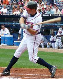 A man in a white baseball uniform with dark red and navy trim and wearing a navy blue baseball helmet swings a baseball bat