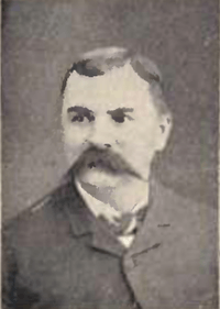 Head of a white man with a wide pointed mustache and neatly combed hair, wearing a dark suit coat and bow tie.