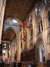 This interior view of Peterborough shows part of the very long nave and the chancel with no screen dividing the building. The structure is mostly of a unified Norman appearance with three levels of simple round-topped arches, only the tall arches of the central tower being Gothic. Other visible features are the ancient painted ceiling, the medieval choir stalls and the modern Rood, which has a gilt wooden figure of Christ on a red cross suspended high in front of the tower arch.