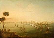 A broad view of a bay. Running vertically from the foreground to the background is a line of 14 anchored ships flying red, white and blue tricolour flags. to their left are four more anchored ships and to the left of these vessels is a distant shoreline. In the foreground of this shore is a hillside on which several men in turbans watch the scene below. To the right of the line a number of ships with all sails set are grouped around the head of the line, as smoke rises from many of the ships on both sides.