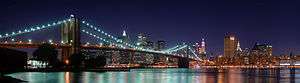 A brightly lit city at night. A suspension bridge lit up with bright blue lights crosses a still body of water in front of the city. The water reflects the blue light, causing rainbow-like stripes to appear on the surface where the lights of the bridge and the city meet.