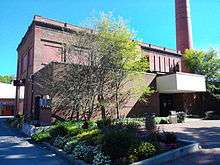 A two-story brick building stands behind a green tree and bed of flowers; in the background, a smokestack rises against a cloudless blue sky.