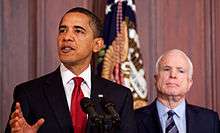 Barack Obama speaking in foreground at an indoor event with an American flag in background; John McCain behind him, somewhat of focus