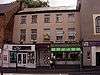 A cream, rendered brick building on three storeys, in a terrace, seen from the north. The lower floor of the building is covered by shop fronts.