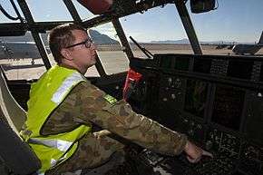 Bespectacled man in camouflage uniform with fluorescent jacket in cockpit of military aircraft