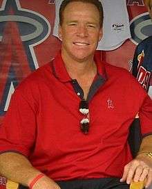 A middle-aged man sitting in a chair and wearing a red polo shirt with a small stylized "A" over the left breast