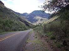 RM 1832, a two-lane road, winds through a canyon with steep hillsides on both sides of the road. In the distance are rugged mountains with steep cliffs covered with desert grasses and brush.