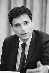 Earnest young man in coat, tie and unruly dark hair, speaking and gesturing