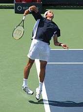 Raonic with both feet in the air at the service line, his racquet behind him, and his eyes looking up.