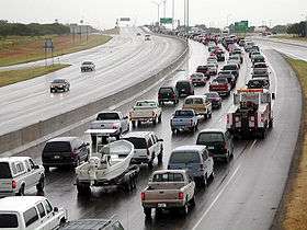 Traffic along a wet highway. Most of the cars are on the right side of the road; however, at least two cars are on the left side.