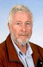 A man with white hair and beard wearing a blue button-down shirt and red coat stands against a sky blue background.