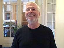 Head and shoulders of a man in his sixties with close-cropped white hair and a short beard, wearing a black T-shirt.