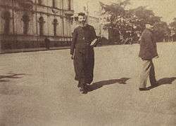 A faded snapshot of Carr Smith walking down a wide street wearing a black cassock and cap.