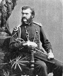 A monochrome photograph portrait of a man with light skin and dark hair, including a bushy muttonchop whiskers and moustache, the man seated, holding a thin sword at rest on his leg, wearing a military uniform decorated with epaulets, braid and buttons