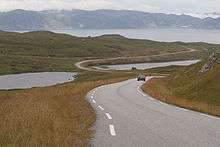 A road runs through a grassland tundra with the ocean in the background