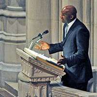 A man, wearing a black suit and white shirt, is standing on a podium while giving a speech.